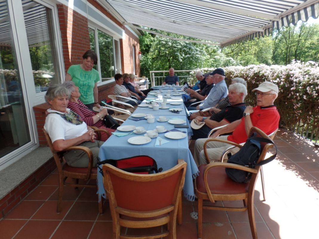 Personen sitzen an einer Kaffeetafel auf einer schattigen Terrasse.